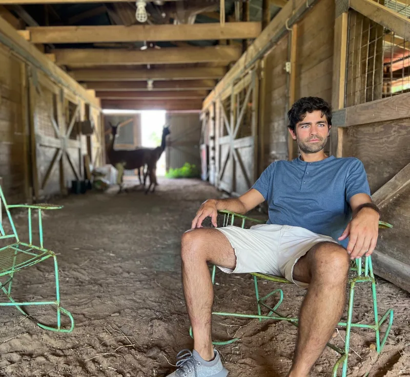 Ο Adrian Grenier κάθεται σε μια καρέκλα μέσα σε ένα αχυρώνα, ενώ δύο αλπακά διακρίνονται στο βάθος.
