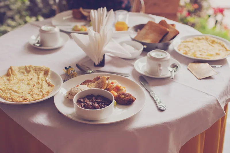 Σερβιρισμένο τραπέζι για κυριακάτικο brunch στο μπαλκόνι με διάφορα πιάτα, φρυγανισμένο ψωμί και λευκά σερβίτσια.