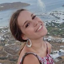 Νεαρή γυναίκα με μακριά μαλλιά και μεγάλα σκουλαρίκια χαμογελάει χαρούμενα με φόντο ένα κυκλαδίτικο τοπίο.
