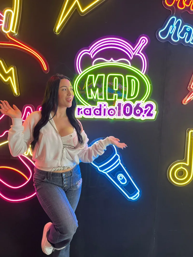 Η Σίλια Δούμου στο Mad Radio 106,2 ποζάρει μπροστά από φωτεινά νέον σημάδια με μικρόφωνο και το λογότυπο του σταθμού.