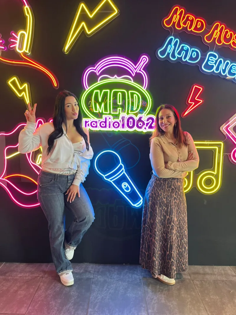 Η Σίλια Δούμου στο Mad Radio 106,2 ποζάρει μπροστά από φώτα νέον με το λογότυπο του σταθμού και ένα μικρόφωνο.
