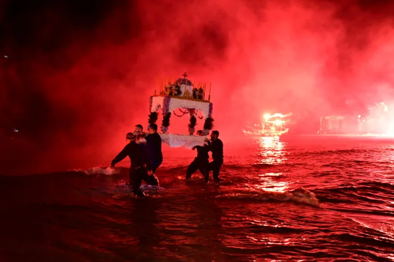Άντρες μεταφέρουν τον Επιτάφιο μέσα στη θάλασσα με κόκκινα πυροτεχνήματα να φωτίζουν τη νύχτα.