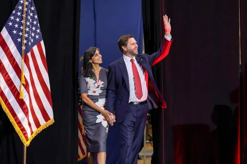 Ο JD Vance και η σύζυγός του Usha Vance χαιρετούν το κοινό δίπλα στην αμερικανική σημαία, σε μια στιγμή πολιτικής σάτιρας.