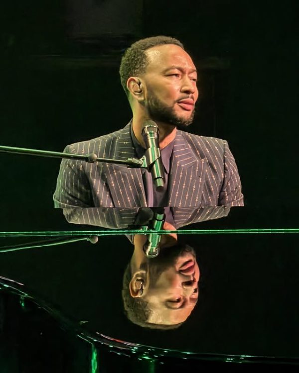 Ο John Legend με ριγέ κοστούμι τραγουδάει στο πιάνο στο Ηρώδειο, αντικατοπτρίζοντας το πρόσωπό του στην γυαλιστερή επιφάνεια.