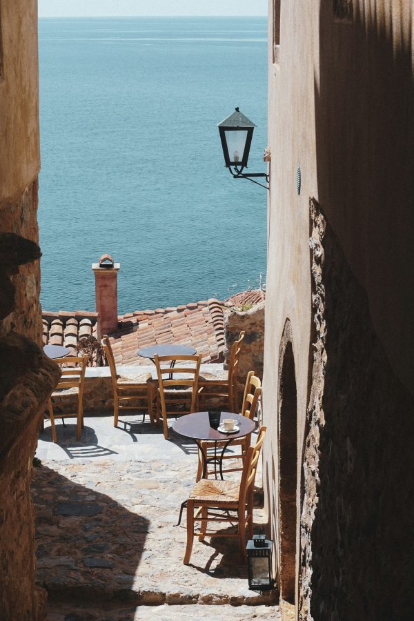 Τραπεζάκια και καρέκλες σε γραφικό σοκάκι με θέα τη θάλασσα, ιδανικός προορισμός για το Πάσχα.