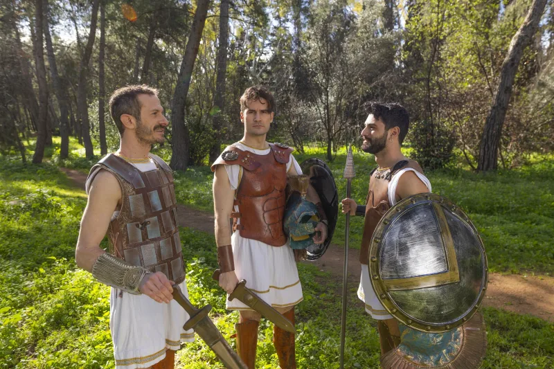 Τρεις άνδρες με αρχαίες στολές πολεμιστών, πανοπλίες και ασπίδες στέκονται σε δασώδη περιοχή, προετοιμαζόμενοι για τον "πόλεμο" της σειράς.