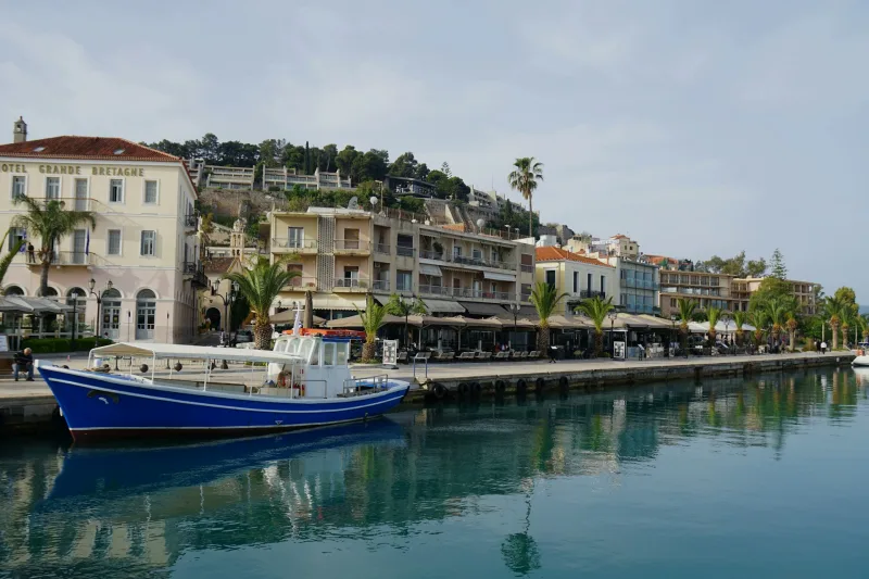 Γαλάζιο καραβάκι αγκυροβολημένο σε λιμάνι με παραθαλάσσια κτίρια και φοίνικες, ιδανικό για ταξίδι με τον σκύλο.
