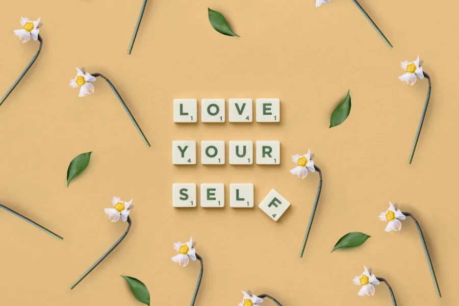 Γράμματα Scrabble γράφουν "LOVE YOUR SELF" ανάμεσα σε λουλούδια και φύλλα, τονίζοντας την αυτοφροντίδα και την προσωπική ανάπτυξη.