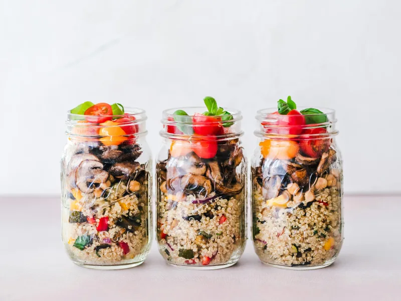 Τρία Mason jars γεμάτα με πολύχρωμες σαλάτες, έτοιμες για το γραφείο, με στρώσεις λαχανικών και κινόα.