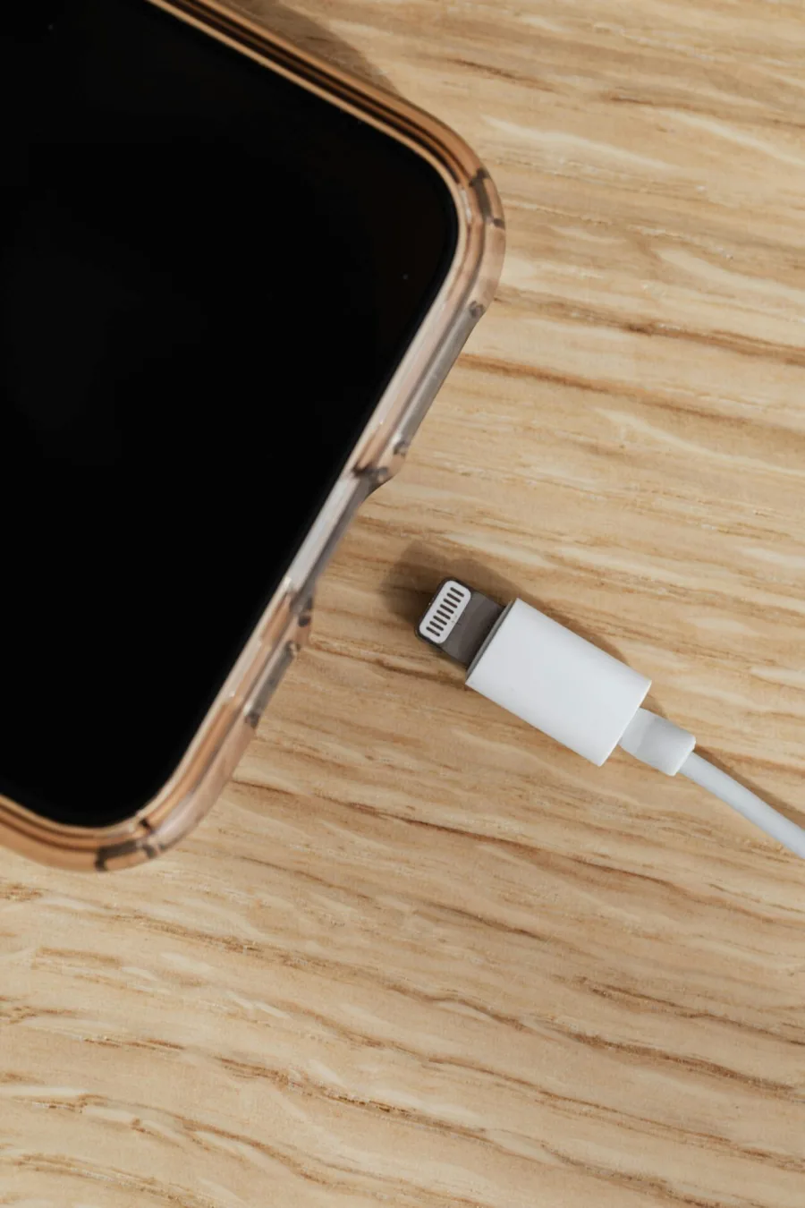 Κοντινό πλάνο ενός smartphone δίπλα σε ένα καλώδιο φόρτισης Lightning πάνω σε ξύλινη επιφάνεια.