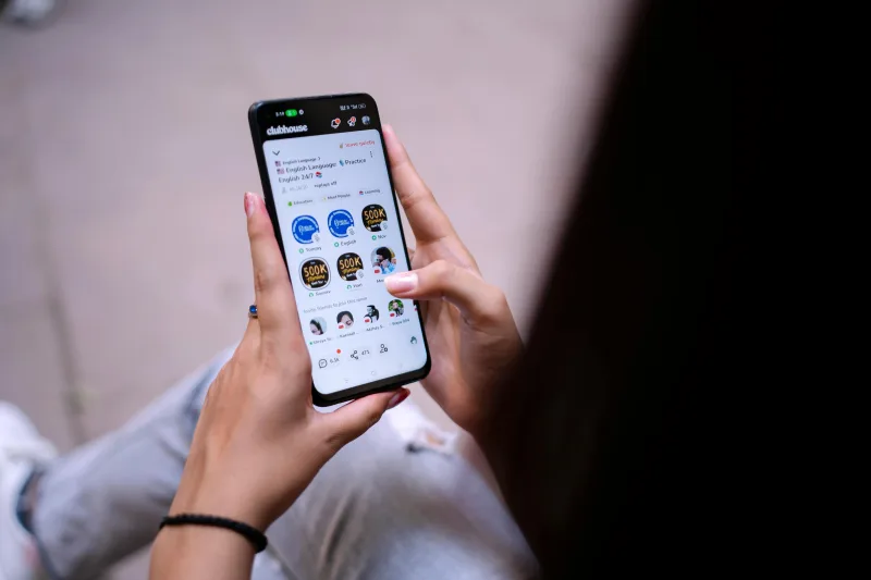 Χέρια που κρατούν smartphone και χρησιμοποιούν την εφαρμογή Clubhouse, εστιάζοντας στα digital red flags που μπορεί να επηρεάσουν μια σχέση.