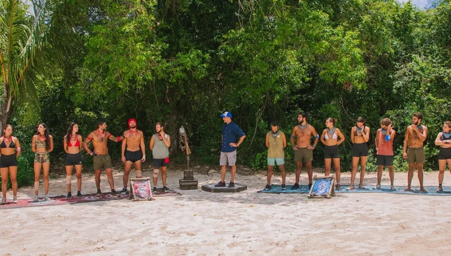 Ομάδα παικτών του Survivor στέκεται σε παραλία με τροπική βλάστηση, περιμένοντας τις εξελίξεις στο παιχνίδι.