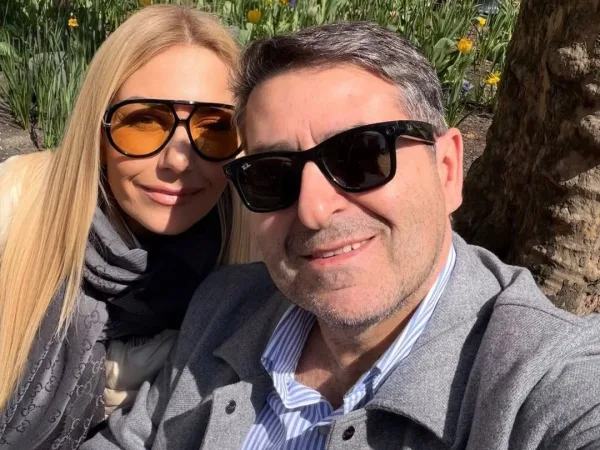Η Τίνα Μεσσαροπούλου και ο Γιώργος Μυλωνάκης ποζάρουν χαμογελαστοί, με φόντο λουλούδια και δέντρο, σε μια ηλιόλουστη μέρα.
