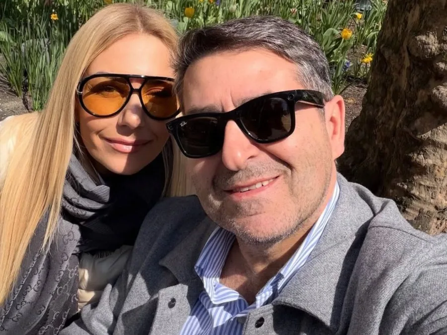 Η Τίνα Μεσσαροπούλου και ο Γιώργος Μυλωνάκης ποζάρουν χαμογελαστοί, με φόντο λουλούδια και δέντρο, σε μια ηλιόλουστη μέρα.