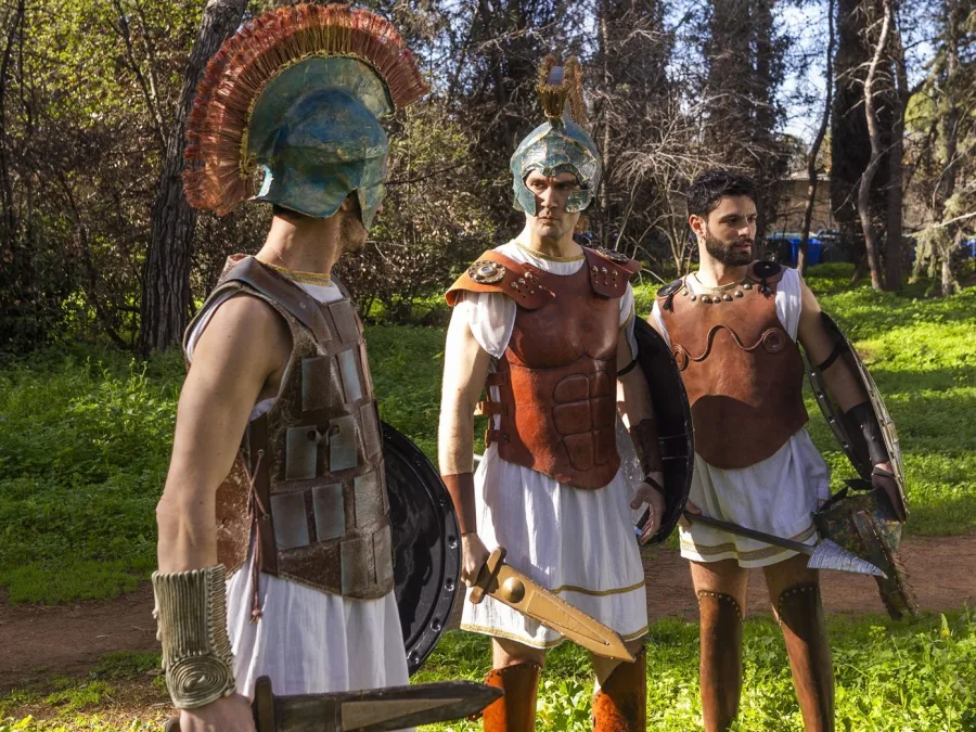 Τρεις άνδρες με αρχαιοελληνικές στολές και όπλα στέκονται σε δασώδη περιοχή, προετοιμαζόμενοι για τον "πόλεμο" της σειράς.