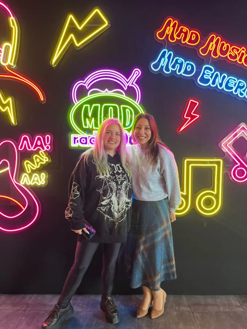 Η Κατερίνα Βλάχου στο Mad Radio 106.2 ποζάρει χαμογελαστή μπροστά από νέον επιγραφές με θέμα τη μουσική και την ενέργεια.