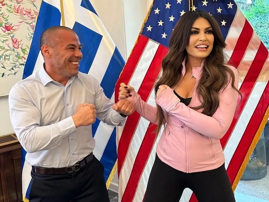 Ο Μιχάλης Ζαμπίδης και η Kimberly Guilfoyle ποζάρουν με υψωμένες γροθιές μπροστά από σημαίες.