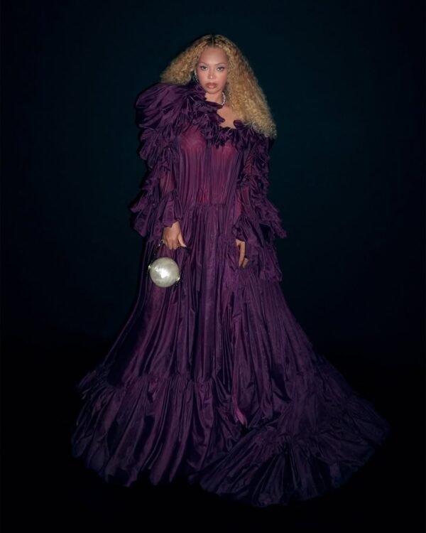 Η Beyoncé με ογκώδες μοβ φόρεμα και σφαιρική τσάντα σε μαύρο φόντο