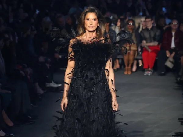 Η Cindy Crawford περπατά σε πασαρέλα φορώντας μαύρο φόρεμα με φτερά.