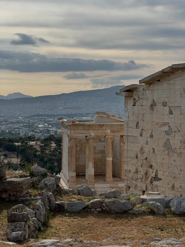 Αρχαίο μνημείο με κολόνες και θέα στην Αθήνα, θυμίζοντας καλοκαίρι στο Αιγαίο.