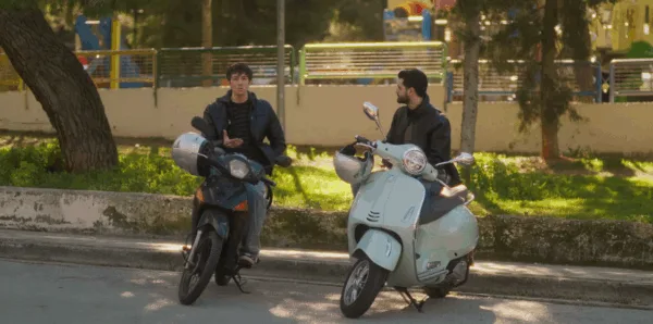 Δύο άνδρες πάνω σε μοτοσικλέτες συνομιλούν σε εξωτερικό χώρο.
