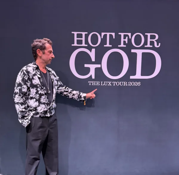 Ο άνδρας δείχνει προς το λογότυπο "HOT FOR GOD THE LUX TOUR 2026" σε φόντο σκούρο μπλε.