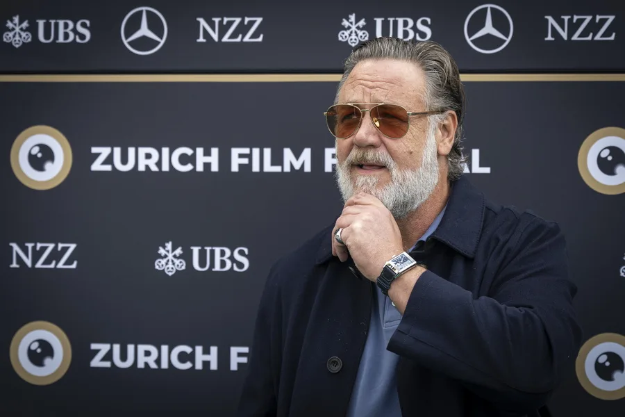 Ο Russell Crowe με γυαλιά ηλίου και γκρι γένια ποζάρει μπροστά από φόντο του Zurich Film Festival.