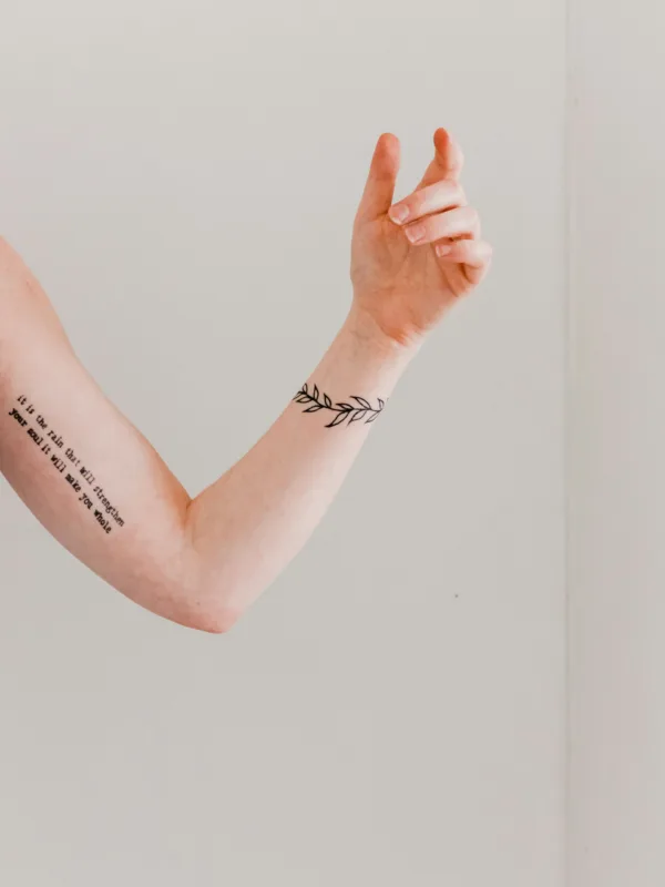 Διακριτικό tattoo με κλαδί ελιάς στον καρπό και φράση στο μπράτσο, ιδανικό για πρώτο tattoo.