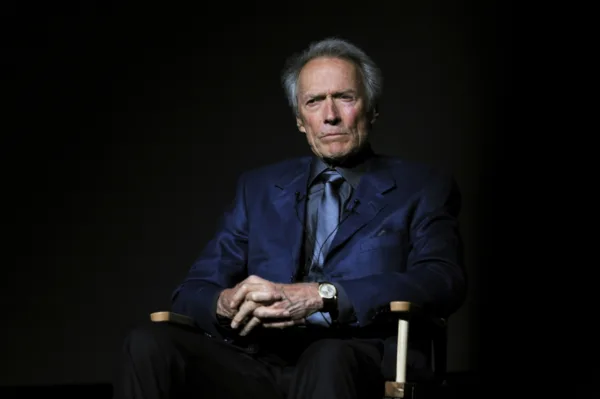 Ο Clint Eastwood κάθεται σε καρέκλα σκηνοθέτη, με το βλέμμα του στραμμένο προς τον φακό, σε μια σκοτεινή ατμόσφαιρα.