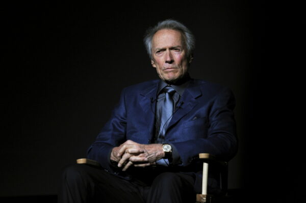 Ο Clint Eastwood κάθεται σε καρέκλα σκηνοθέτη, με το βλέμμα του στραμμένο προς τον φακό, σε μια σκοτεινή ατμόσφαιρα.