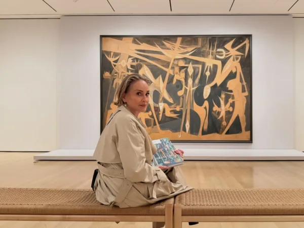 Η Sharon Stone κάθεται σε ένα παγκάκι σε γκαλερί τέχνης, κρατώντας ένα βιβλίο με έργα τέχνης.