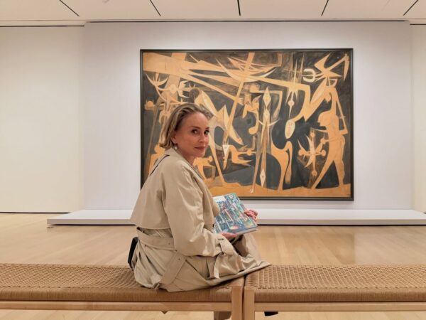 Η Sharon Stone κάθεται σε ένα παγκάκι σε γκαλερί τέχνης, κρατώντας ένα βιβλίο με έργα τέχνης.