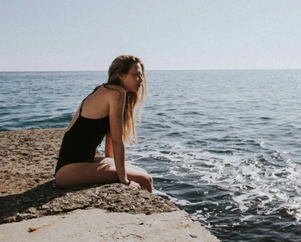 Γυναίκα με μαγιό κάθεται σε βράχο δίπλα στη θάλασσα, απολαμβάνοντας την ηρεμία και τις ταξιδιωτικές εμπειρίες του 2026.