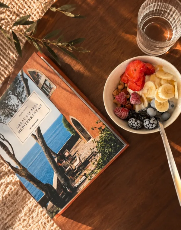 Ένα μπολ με φρούτα και ένα βιβλίο με τίτλο Great Escapes Mediterranean βρίσκονται πάνω σε ξύλινο τραπέζι.