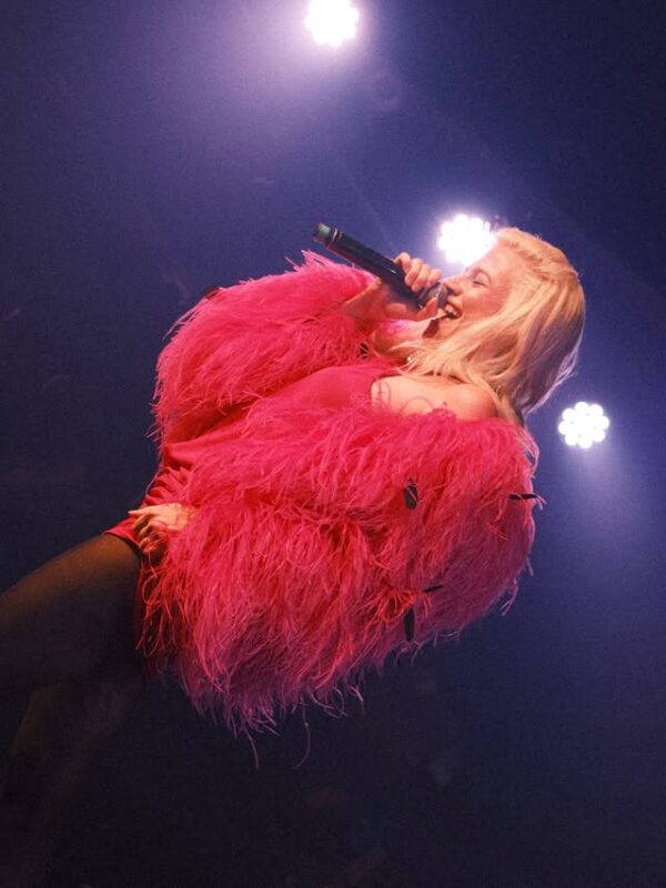 Η Pixie Lott τραγουδάει με φόρεμα φούξια με φτερά, φορώντας μαύρο καλσόν, στη σκηνή.