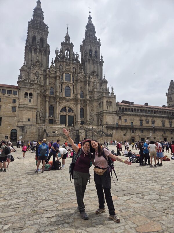 Δύο γυναίκες ολοκληρώνουν το Camino de Santiago μπροστά από τον εντυπωσιακό καθεδρικό ναό, έτοιμες για νέες εμπειρίες.