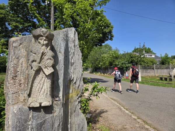Γλυπτό μοναχού στο Camino de Santiago, με δύο πεζοπόρους να βαδίζουν στον δρόμο, σε ένα ταξίδι που αλλάζει τη ζωή.
