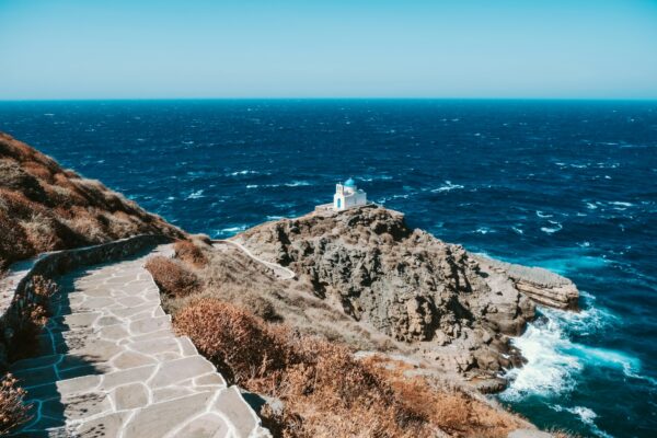 Μια γραφική διαδρομή οδηγεί σε ένα λευκό εκκλησάκι με μπλε τρούλο, πάνω σε βράχια, σε ένα από τα ελληνικά νησιά.