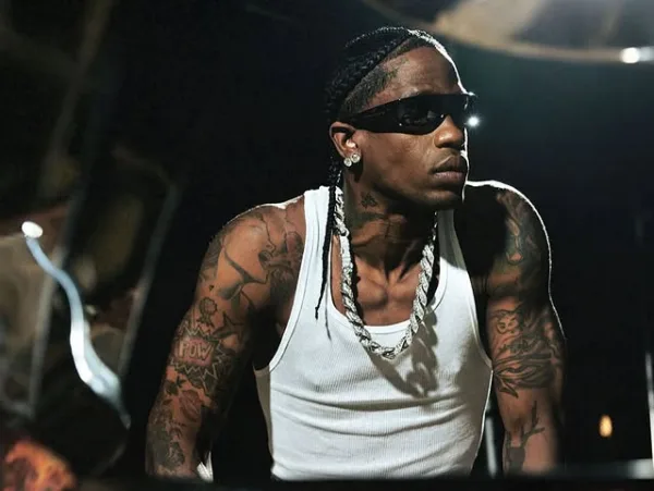 Ο Travis Scott με γυαλιά ηλίου και ασημένια αλυσίδα, φορώντας λευκό φανελάκι, παρουσιάζει τα τατουάζ του.