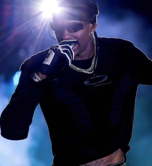 Ο Travis Scott τραγουδάει με γάντια και κοσμήματα, ενώ φωτίζεται από έντονο φως στη σκηνή.