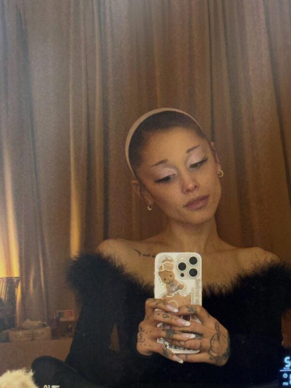 Η Ariana Grande τραβάει selfie με το κινητό της, φορώντας ένα μαύρο φόρεμα με φτερά, λίγο πριν παγιδευτεί σε ασανσέρ.