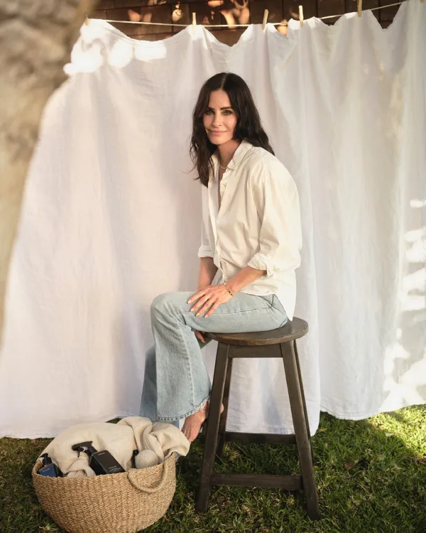 Η Courtney Cox ποζάρει καθισμένη σε σκαμπό, με φόντο λευκό ύφασμα, ενώ δίπλα της βρίσκεται ένα καλάθι με ρούχα.