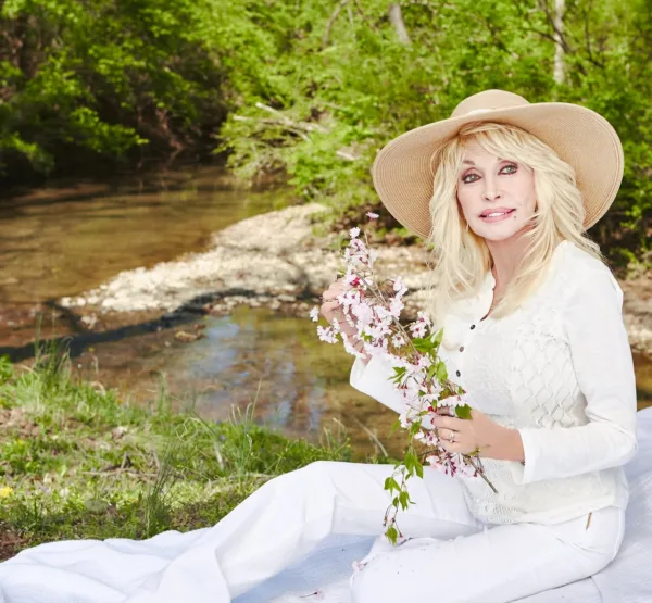 Η Dolly Parton με καπέλο και λουλούδια δίπλα σε ένα ποταμάκι, λίγο πριν φύγει.
