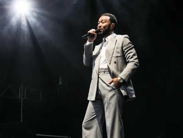 Ο John Legend τραγουδάει με μικρόφωνο στη σκηνή, φορώντας κοστούμι, στο πλαίσιο της συνεργασίας του με τον Pharrell Williams για τον νέο ήχο του «Muse».