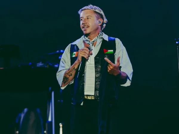 Ο Macklemore τραγουδάει στο stage, φορώντας γιλέκο με σημαίες, κατά την περιοδεία του.