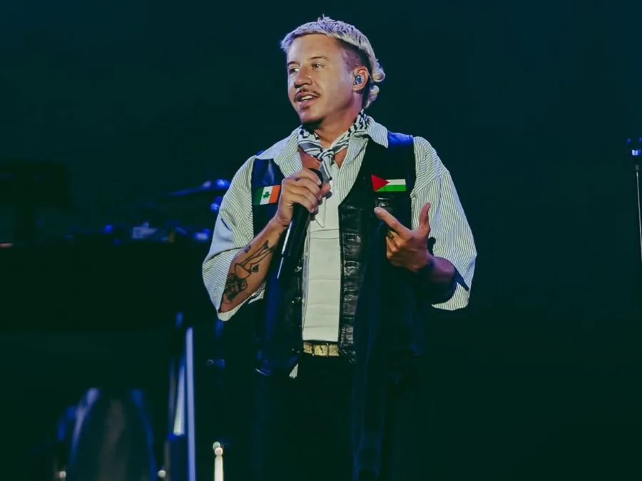 Ο Macklemore τραγουδάει στο stage, φορώντας γιλέκο με σημαίες, κατά την περιοδεία του.