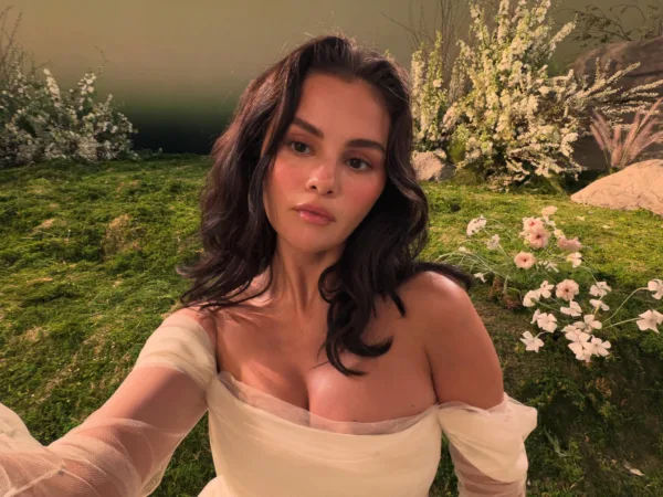 Η Selena Gomez ποζάρει με λευκό φόρεμα σε ένα σκηνικό με πράσινο γκαζόν και λουλούδια.