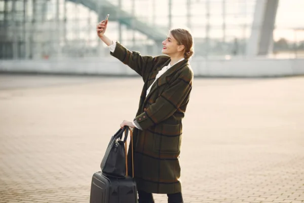 Γυναίκα με βαλίτσες και τσάντα παίρνει selfie έξω από αεροδρόμιο, ιδανική για solo ταξίδι.