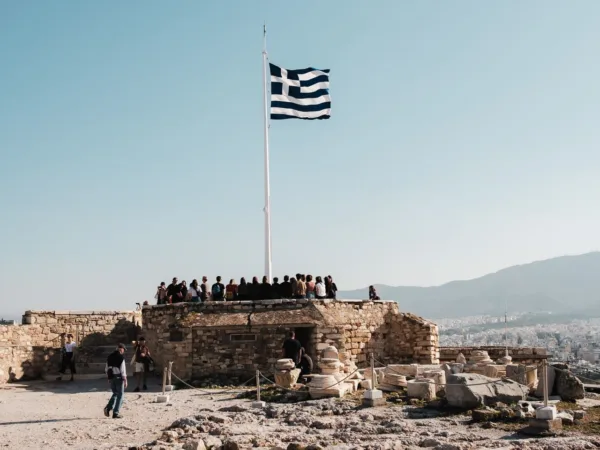 Η ελληνική σημαία κυματίζει στον ιστό πάνω από αρχαιολογικό χώρο με επισκέπτες, τιμώντας την επέτειο της 28ης Οκτωβρίου.