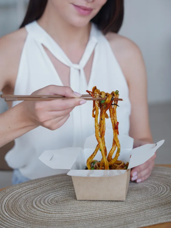 Γυναίκα τρώει noodles από πακέτο με ξυλάκια, εξηγώντας γιατί το σώμα ζητά φαγητό όταν αγχώνεται.